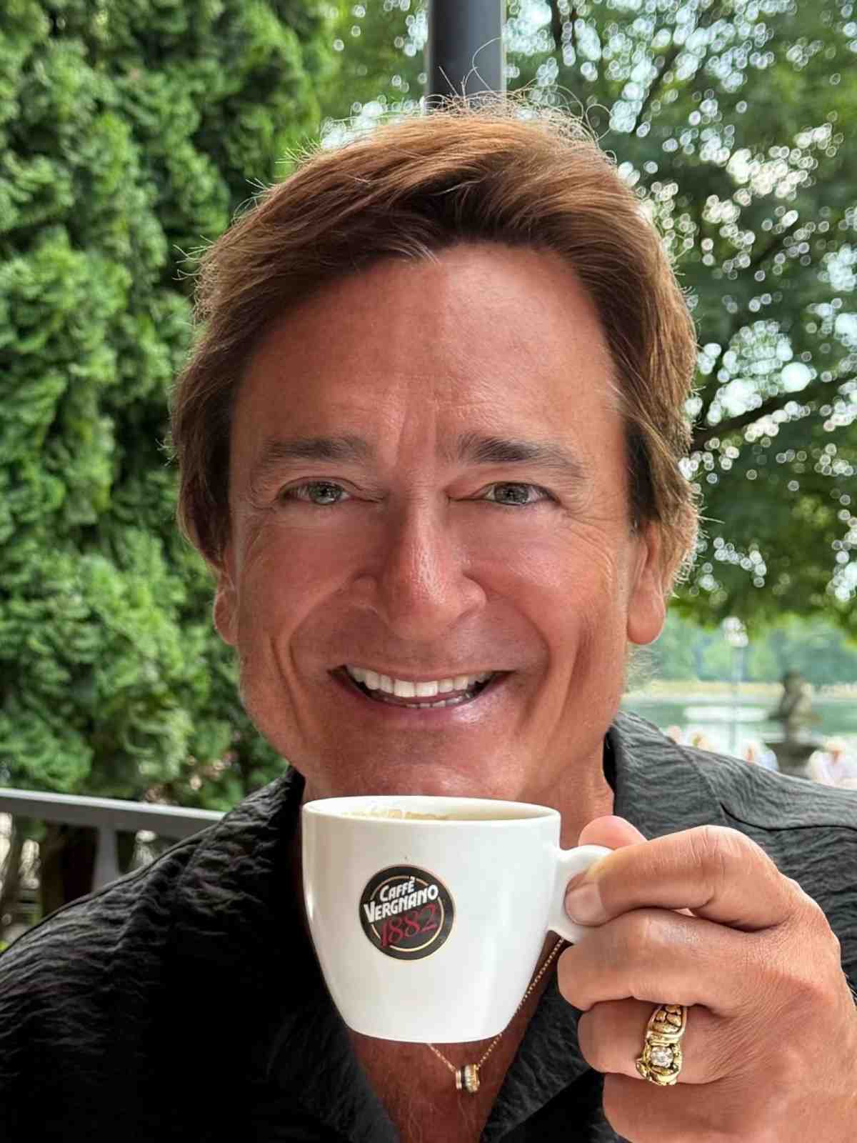 Person smiling, holding a coffee cup outdoors.